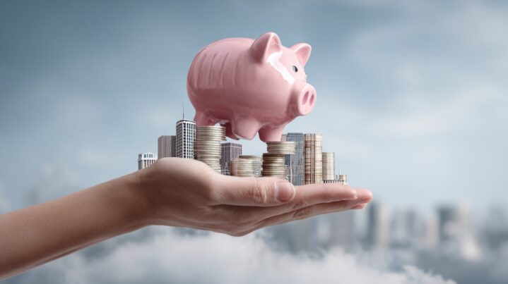 A hand holding a piggy bank above stacks of coins, with an illustration of skyscrapers and business buildings, symbolizing the financial burden of healthcare costs